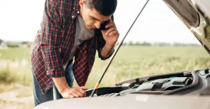 Young man standing next to his car with the hood open, talking on the phone for help, representing frustration and the common issue of a car that won’t start.