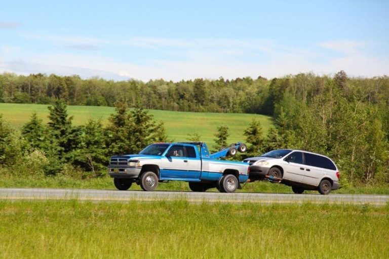 Blue and silver tow truck using a tow dolly to transport a disabled minivan along a scenic countryside highway with green fields and trees in the background