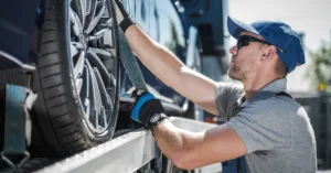 Man in a blue cap and gloves securing a car tire on a transport trailer.