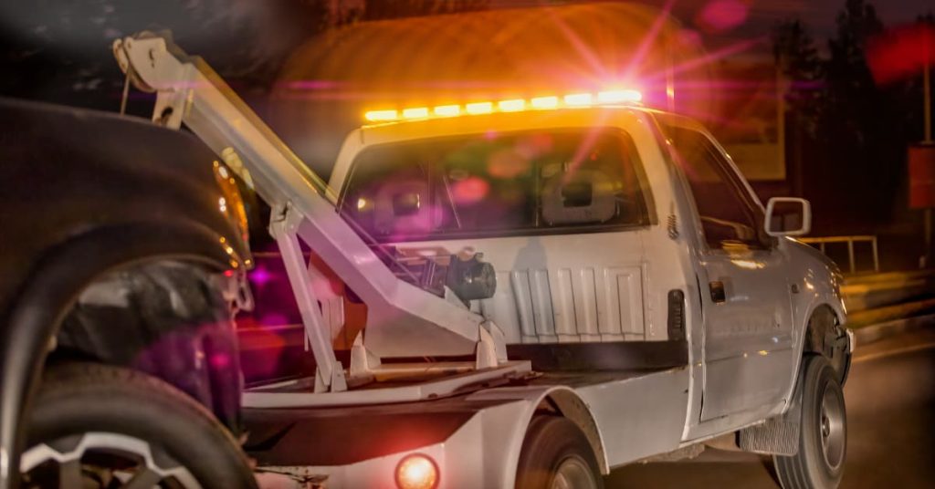 White tow truck with flashing amber lights towing a disabled vehicle on the roadside at night, delivering emergency roadside assistance in low-light conditions
