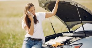 Woman standing by her broken-down car with the hood open, calling for emergency roadside assistance on her phone after experiencing vehicle trouble on the road