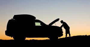 Silhouette of a person checking under the hood of a car at sunset, waiting for 24/7 roadside assistance during a breakdown.