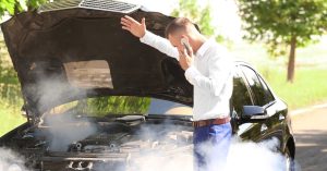 Man standing beside a black car with the hood open, calling for help as smoke rises from the engine, indicating overheating in hot weather.