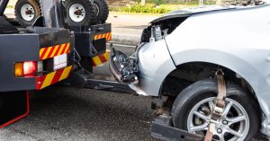 Close-up of a tow truck hauling a silver car with front-end damage, showing the vehicle secured with straps after a roadway collision.