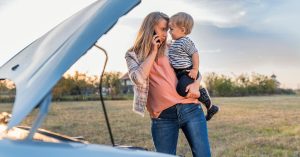 A woman holding a young child while making a phone call next to her broken-down car with the hood open, seeking roadside assistance.