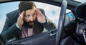 Stressed man in a suit looking through a car window with hands on his head, realizing he is locked out of his vehicle with the keys inside.